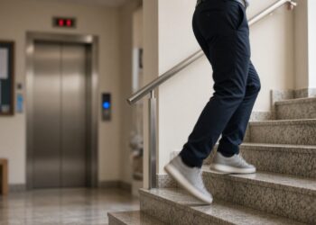 Pessoa subindo a escada de granito no hall de um prédio, com a porta do elevador fechada ao fundo